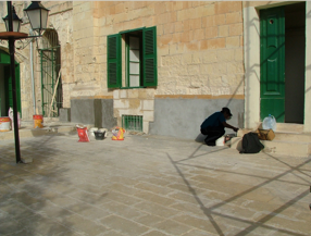 Person crouched near a wall on a cobblestone street with green door and window shutters, surrounded by buckets and bags.