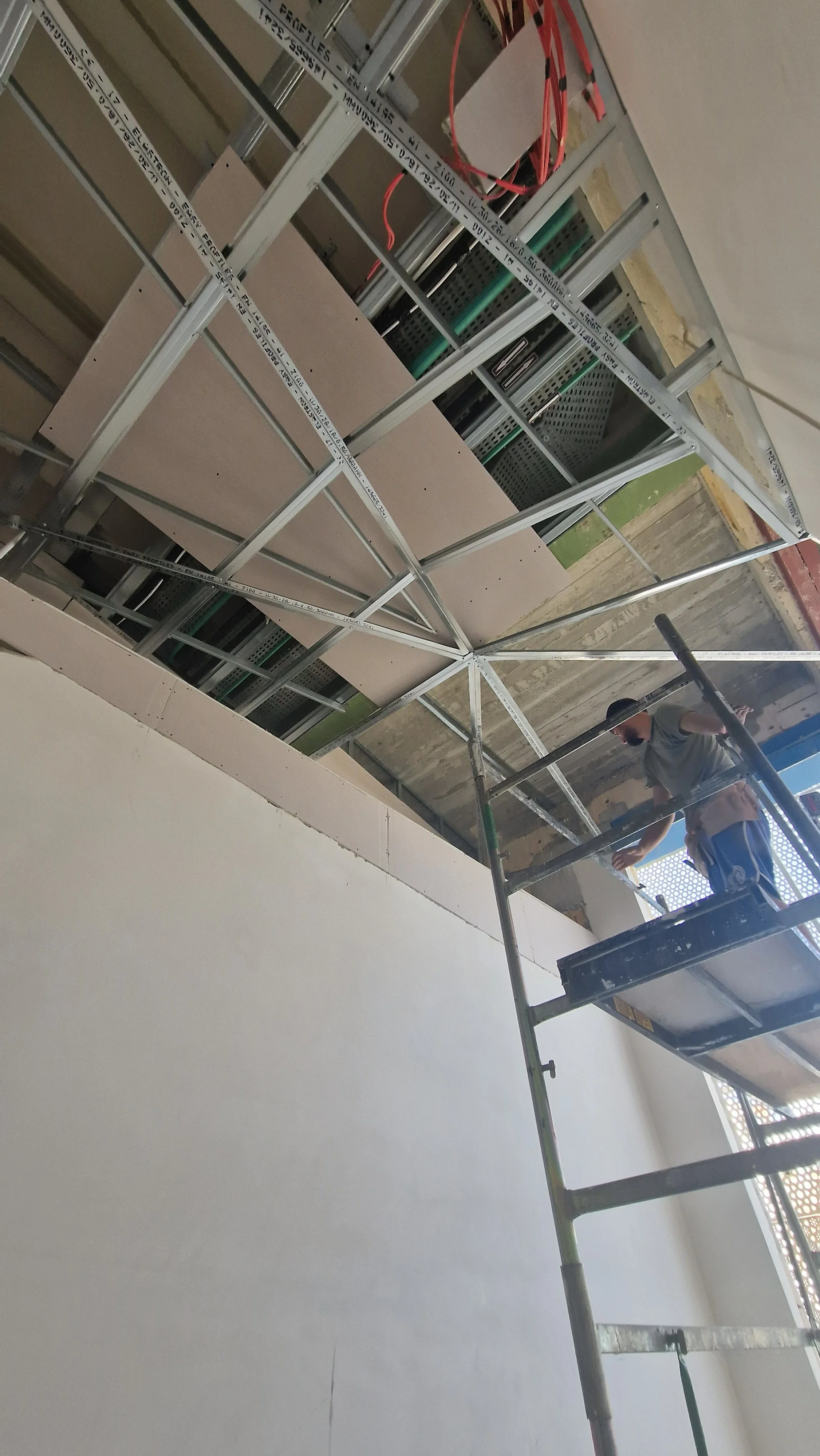 A construction worker is standing on scaffolding, working on a ceiling with exposed metal framing and electrical wiring.
