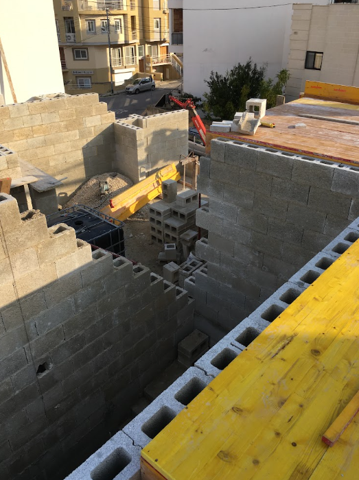 Construction site with gray concrete blocks being assembled into walls, wooden planks, and construction tools. A red mini excavator is visible in the background, with a residential building and street nearby.