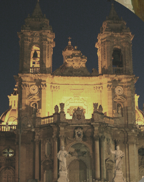 A historic cathedral illuminated at night, with two bell towers, statues in front, and intricate architectural details.