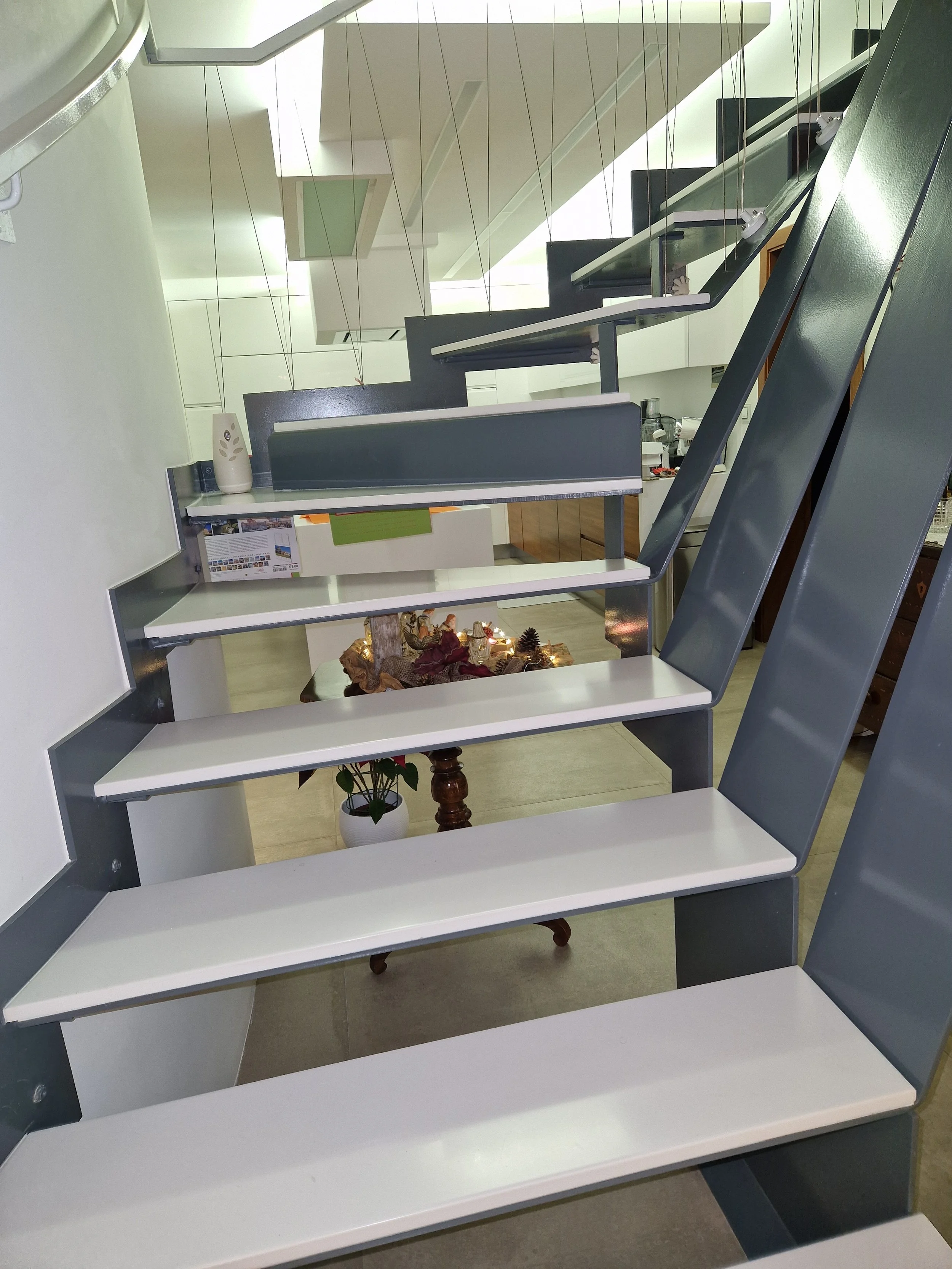A staircase with alternating white and dark gray steps, metal supports, and cable railings indoors. A potted plant and decorative items are visible at the bottom of the staircase.