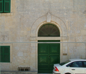 Stone building with green door and shutters, arched window, and a white car parked in front.