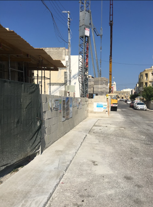 Construction site with a partially built structure, construction materials, and a nearby street with parked cars, under a clear blue sky.