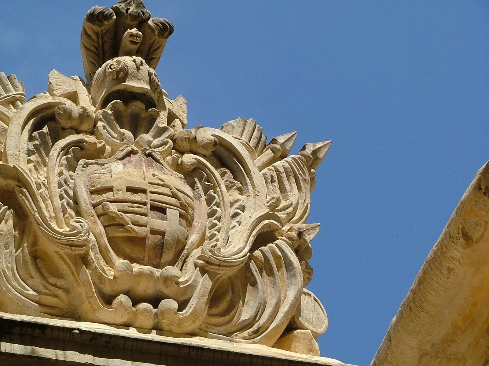 Close-up of an ornate stone crest with a shield, crown, and intricate carvings, set against a blue sky.