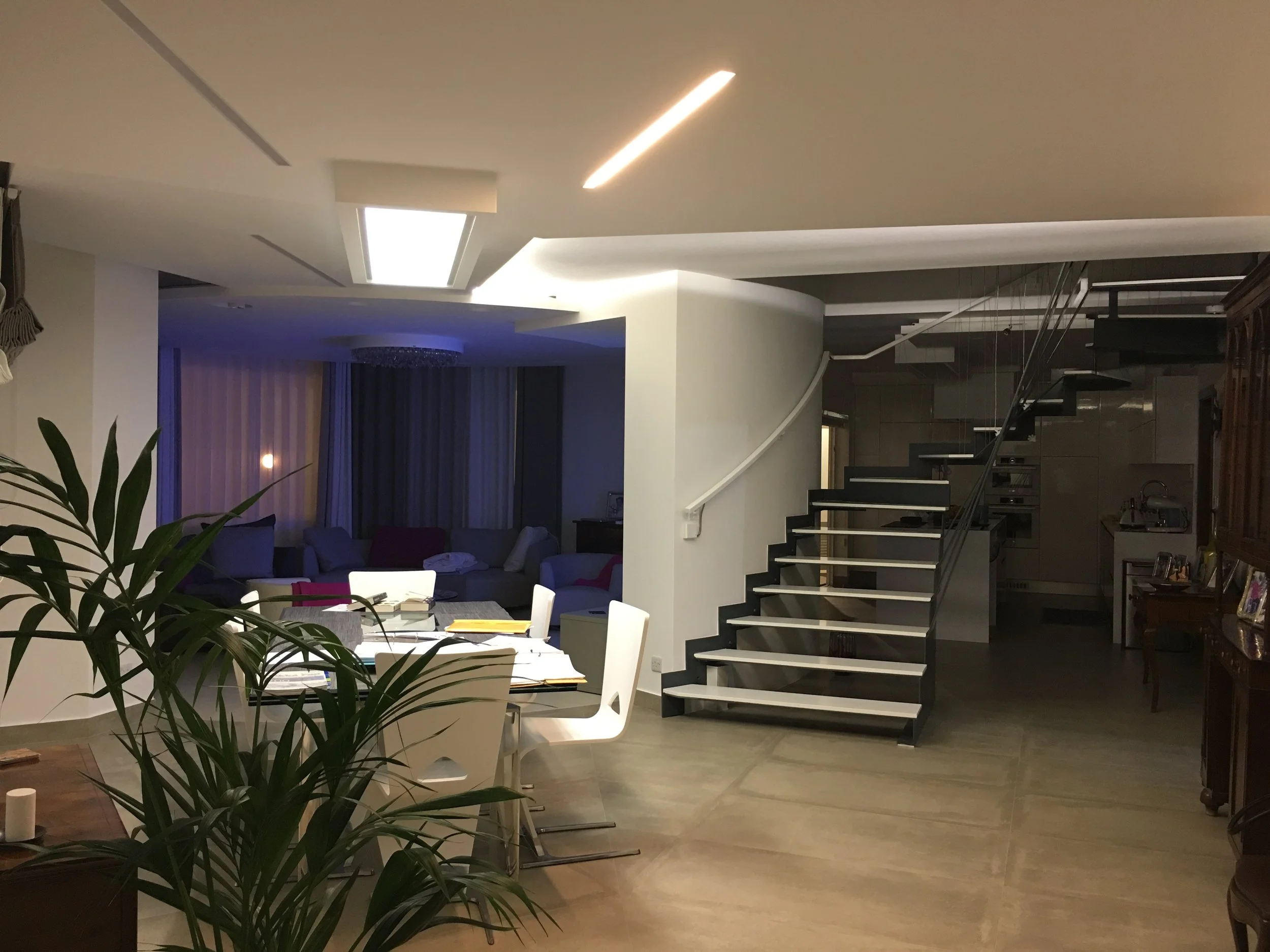 Interior of a modern house with a staircase, dining area with white chairs, and living room with couches. The ceiling has recessed lighting and a skylight. Plants are visible in the foreground.