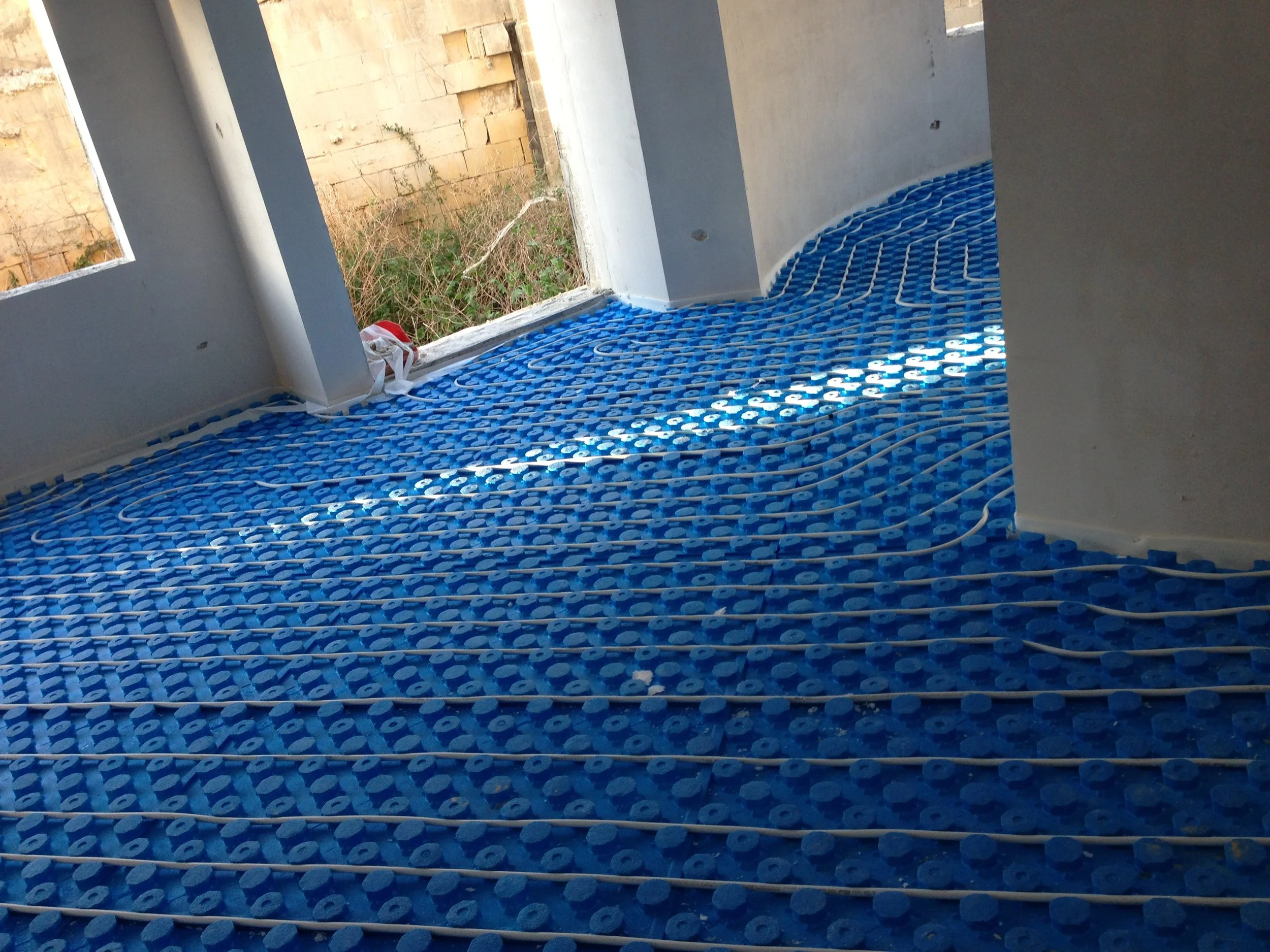 Interior view of a room under construction, showing a radiant floor heating system with blue plastic tubing laid in a patterned grid and white wires, near an unfinished window opening with outdoor vegetation and a brick wall in the background.