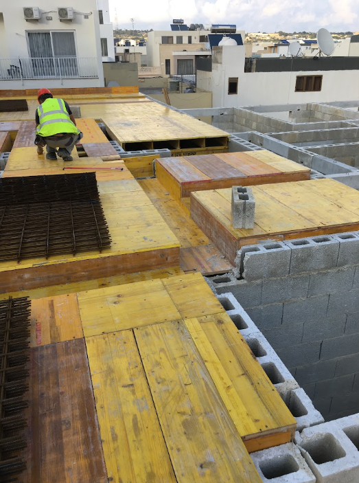 A construction worker wearing a safety helmet and vest crouches on a plywood platform on a building roof under construction, with city buildings and satellite dishes in the background.