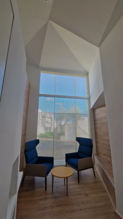 Two blue armchairs and a small round wooden table in a modern room with a large window, wood-paneled walls, and a high, geometric ceiling.