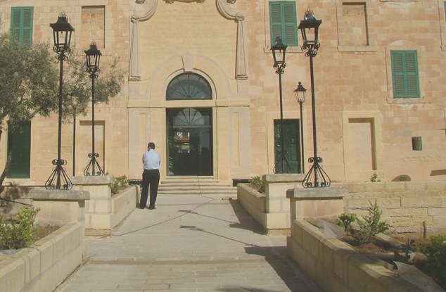 A man in a white shirt and dark pants standing outside a large stone building with green window shutters, arched entrance, and four black street lamps in front.