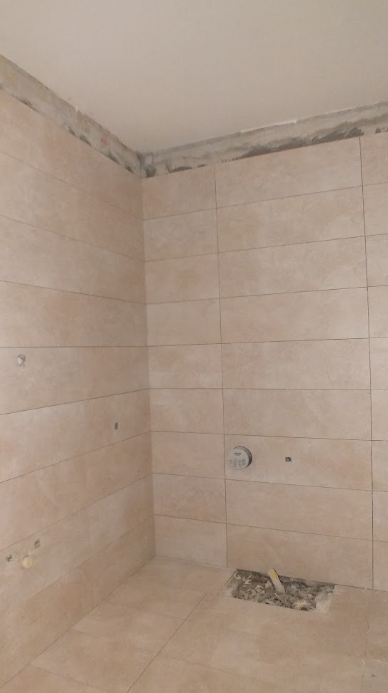 A partially finished bathroom with beige tile walls and floor, with exposed plumbing and an unfinished ceiling.