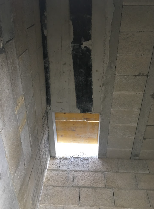 Photo of a small rectangular opening at the bottom of a wall with light coming through, framed with wood.