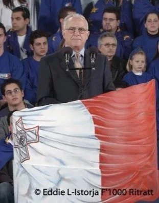 An older man with glasses standing behind a microphone, holding a large French flag. Several people in blue clothing are seen in the background, including children and young adults.