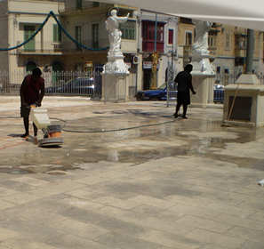 Two workers cleaning or performing maintenance on a paved outdoor area with statues and buildings in the background.