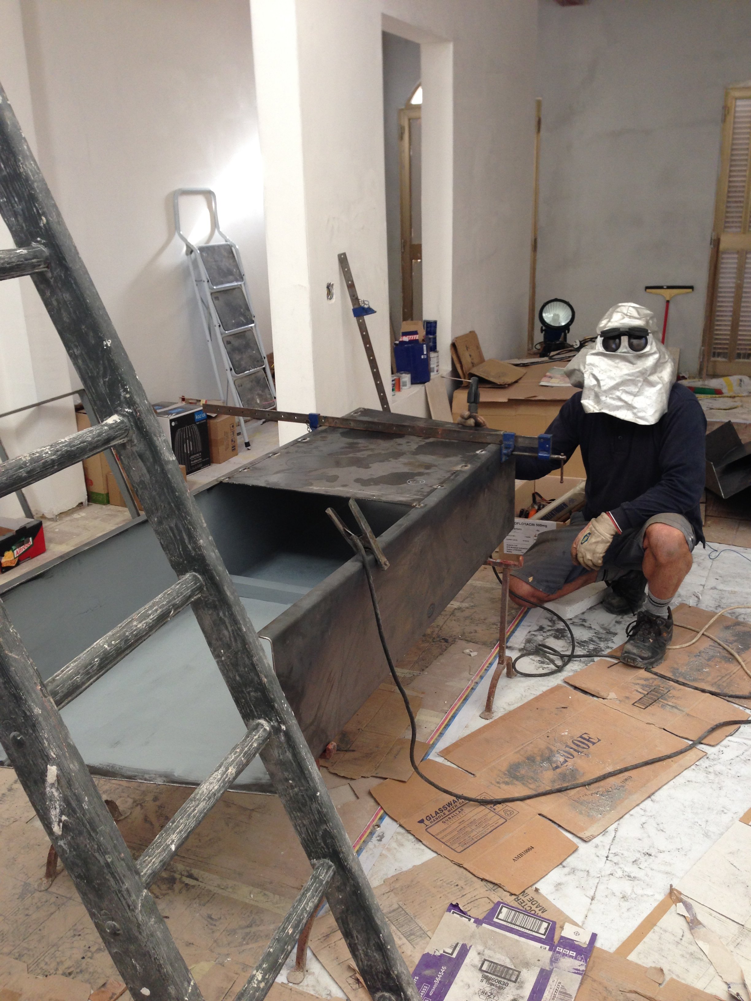 Person wearing protective gear working on construction inside a house, with ladders, tools, and cardboard on the floor.