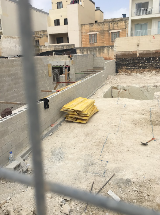 Construction site with a partially built concrete wall, yellow insulation or foam boards stacked on the ground, and residential buildings in the background.
