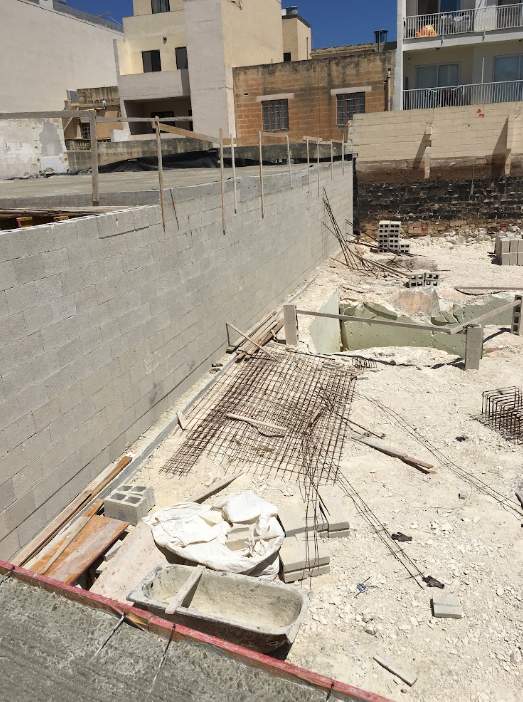 Construction site with concrete blocks, rebar, and building materials, adjacent to existing buildings.