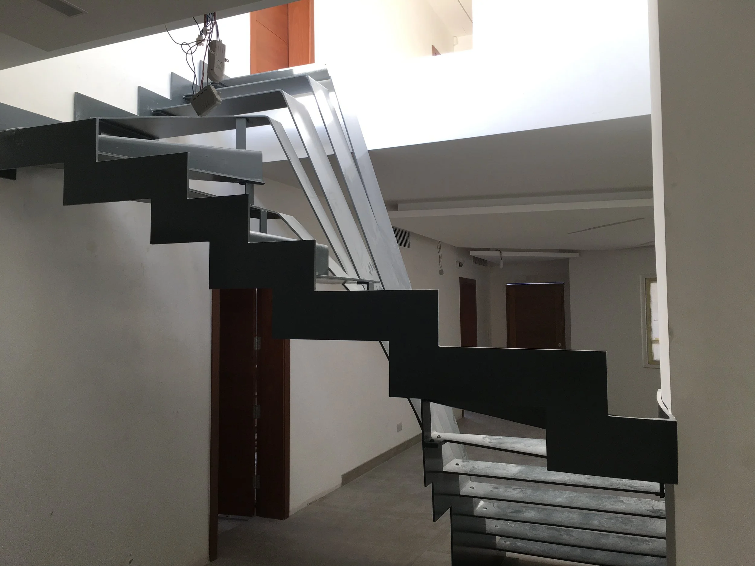 Interior view of a staircase with black steps and metal railings, next to a wall with gray and white walls and a window in the background.