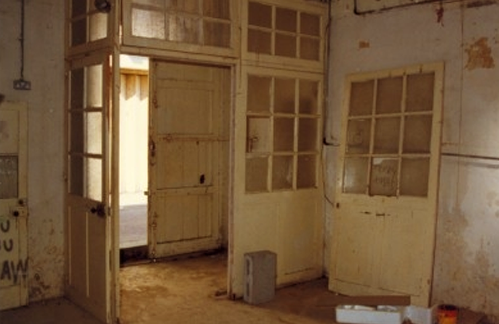 Interior of an old, abandoned building with peeling paint, broken windows, and a doorway leading outside.