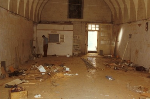 A person standing in a dirty, cluttered room with scattered debris, damaged walls, and a high arched ceiling.
