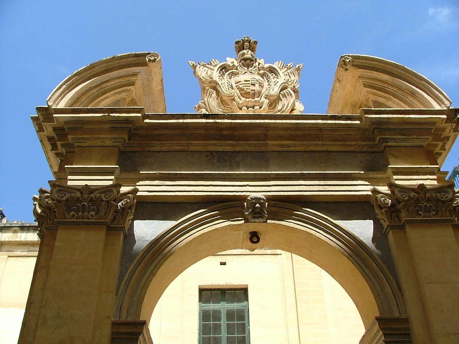 Detail of an ornate stone archway at a historic building, featuring intricate carvings, a decorative crest, and a semi-circular decorative element on top, with a blue sky in the background.