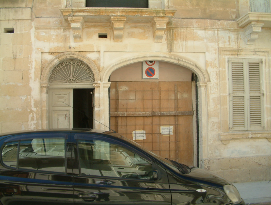 A black car parked in front of an old, weathered stone building with an arched doorway and a closed window with shutters, and a 'no parking' sign above the doorway.