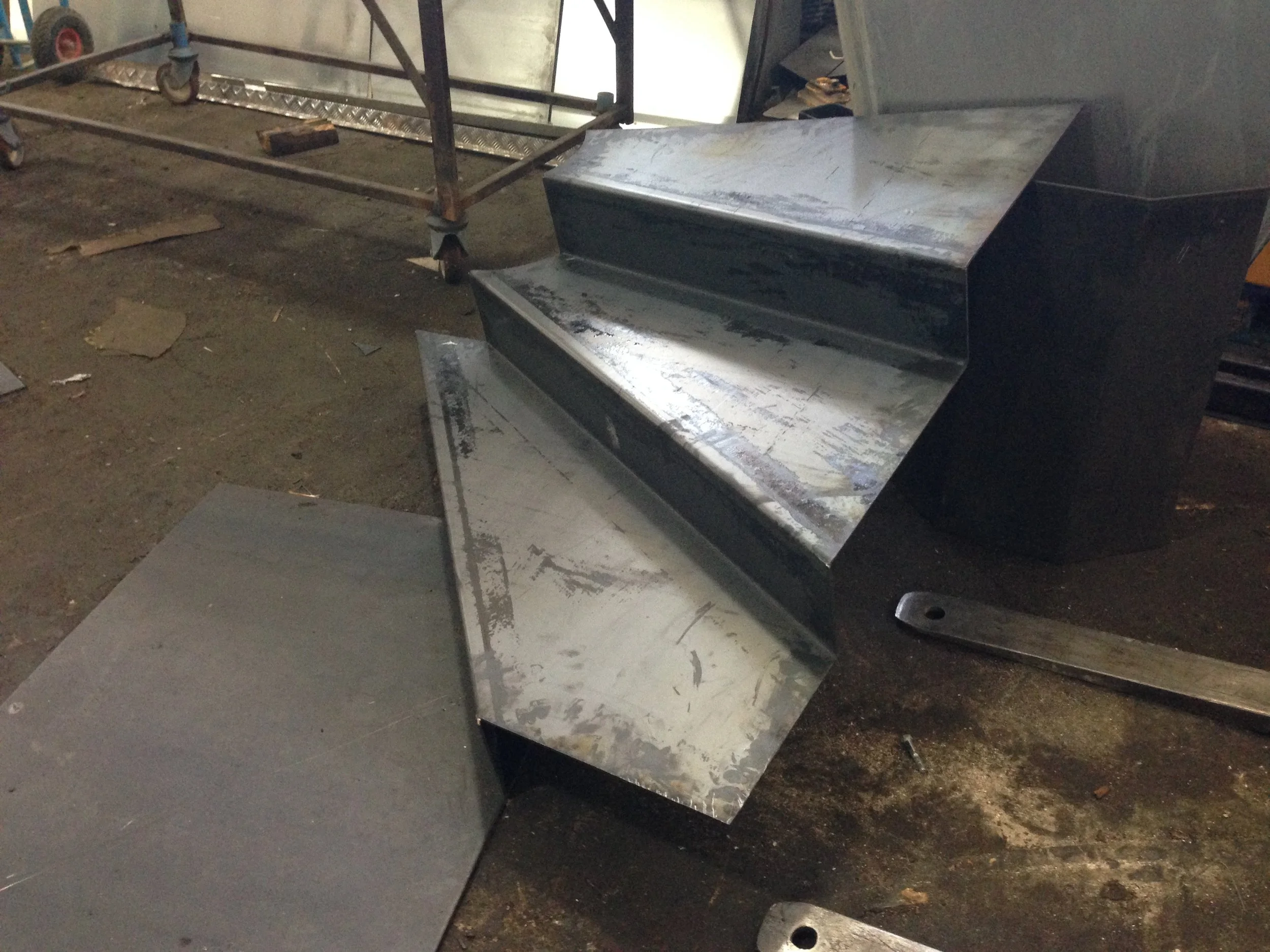Metal equipment, possibly a commercial oven or grill, with a large rectangular opening, located in a dusty, industrial or workshop setting. There is a metal cart with wheels in the background.