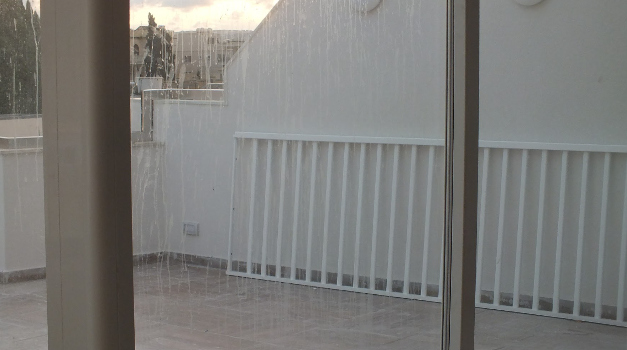 A sliding glass door with raindrops on the glass, looking onto a balcony with a white metal railing and a tiled floor, with buildings visible in the background.