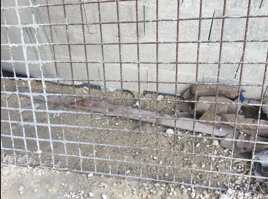 A construction site with a metal grid fence in front of a cinder block wall, dirt and gravel ground, and debris including a paper bag and wooden board.
