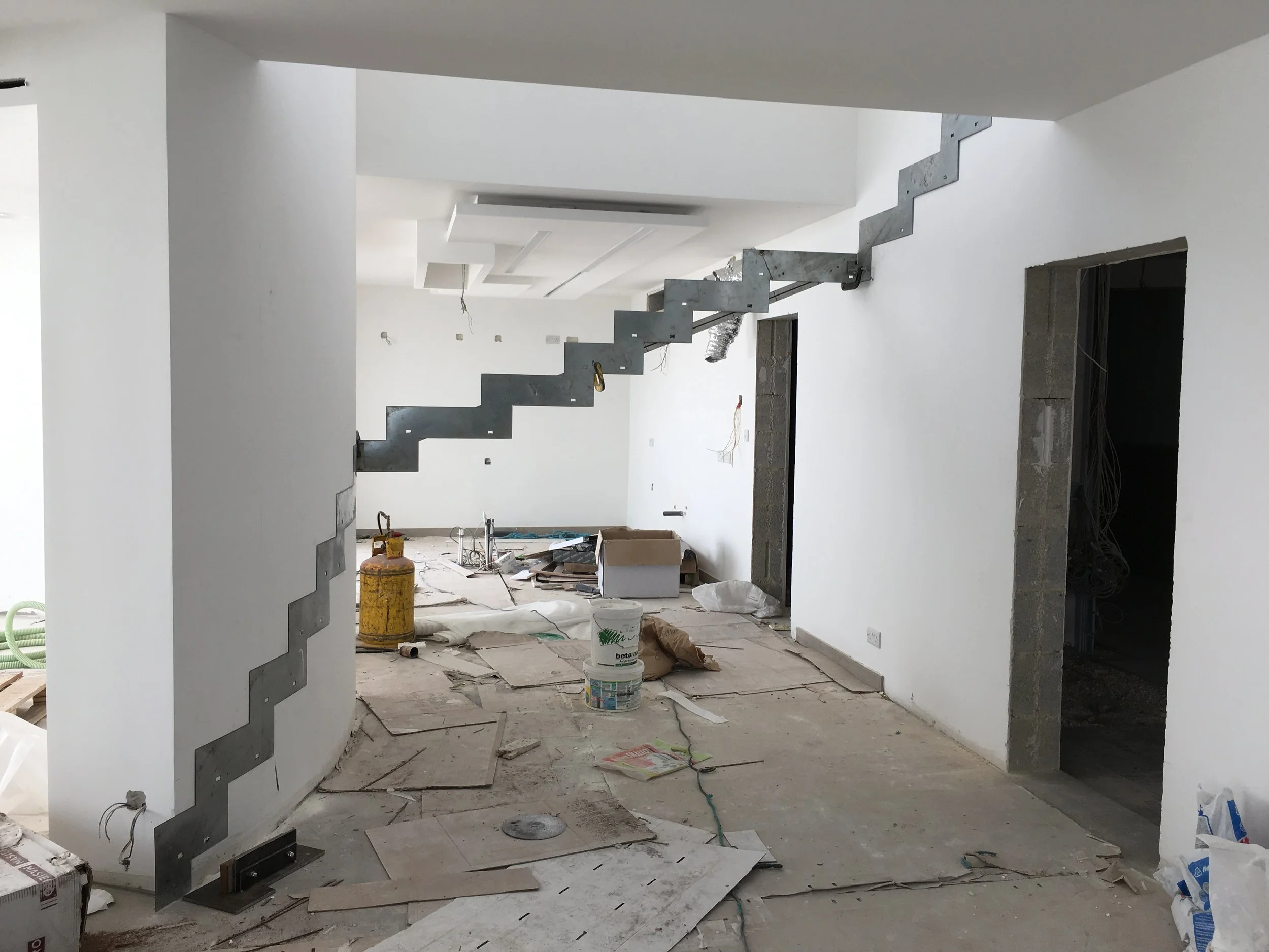 Interior of a house under construction with white walls, an unfinished staircase, construction materials, scattered tools, and debris on the floor.