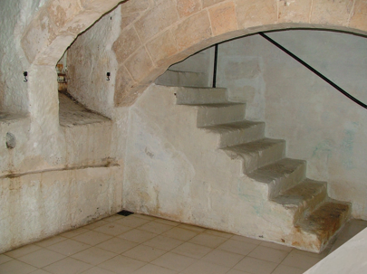 A stone staircase with six steps leading up, with a metal railing on the right side, inside a building with stone and plaster walls.