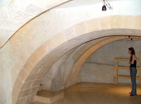 A woman standing inside a large, curved architectural structure made of beige stone, with wooden railing and soft lighting.