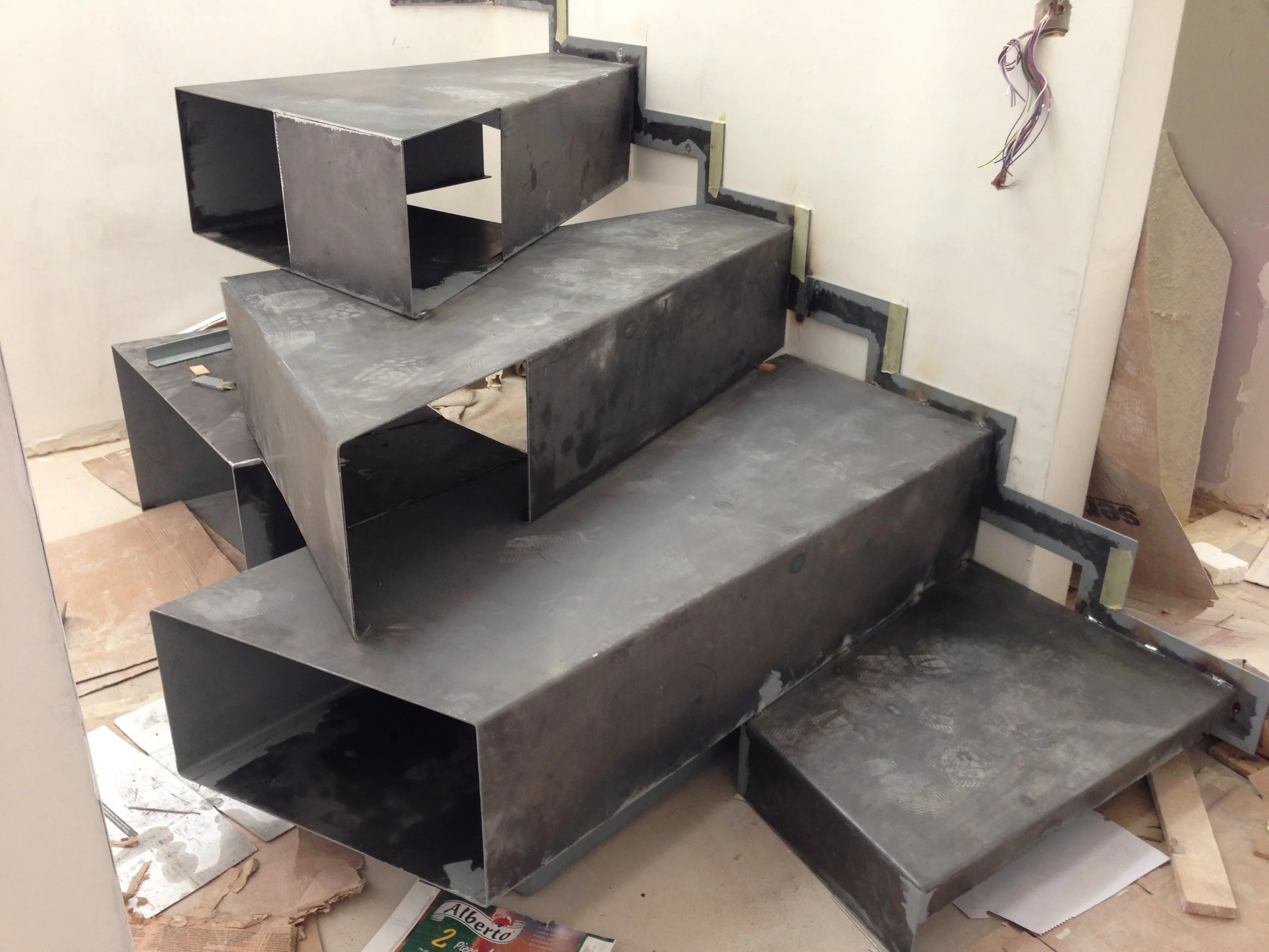 Metal staircase structure in progress, with unpainted, unfinished metal steps and framework, construction materials, and wiring on the wall nearby.