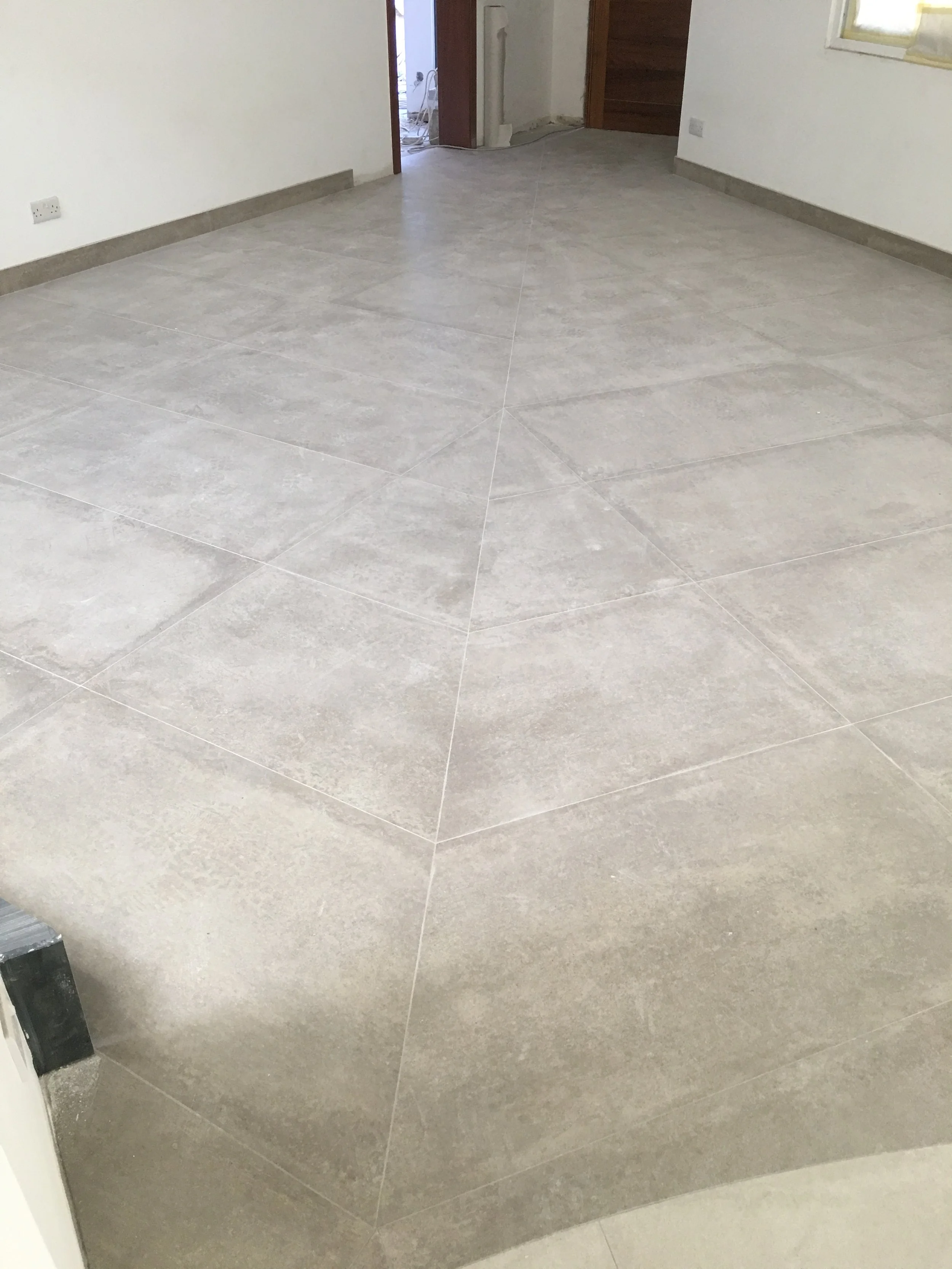 A room with newly installed light gray tiled flooring, white walls, and a wooden door. A window is visible on the right side, and there is an open doorway leading to another room at the back. A black object is partially visible in the bottom left cor