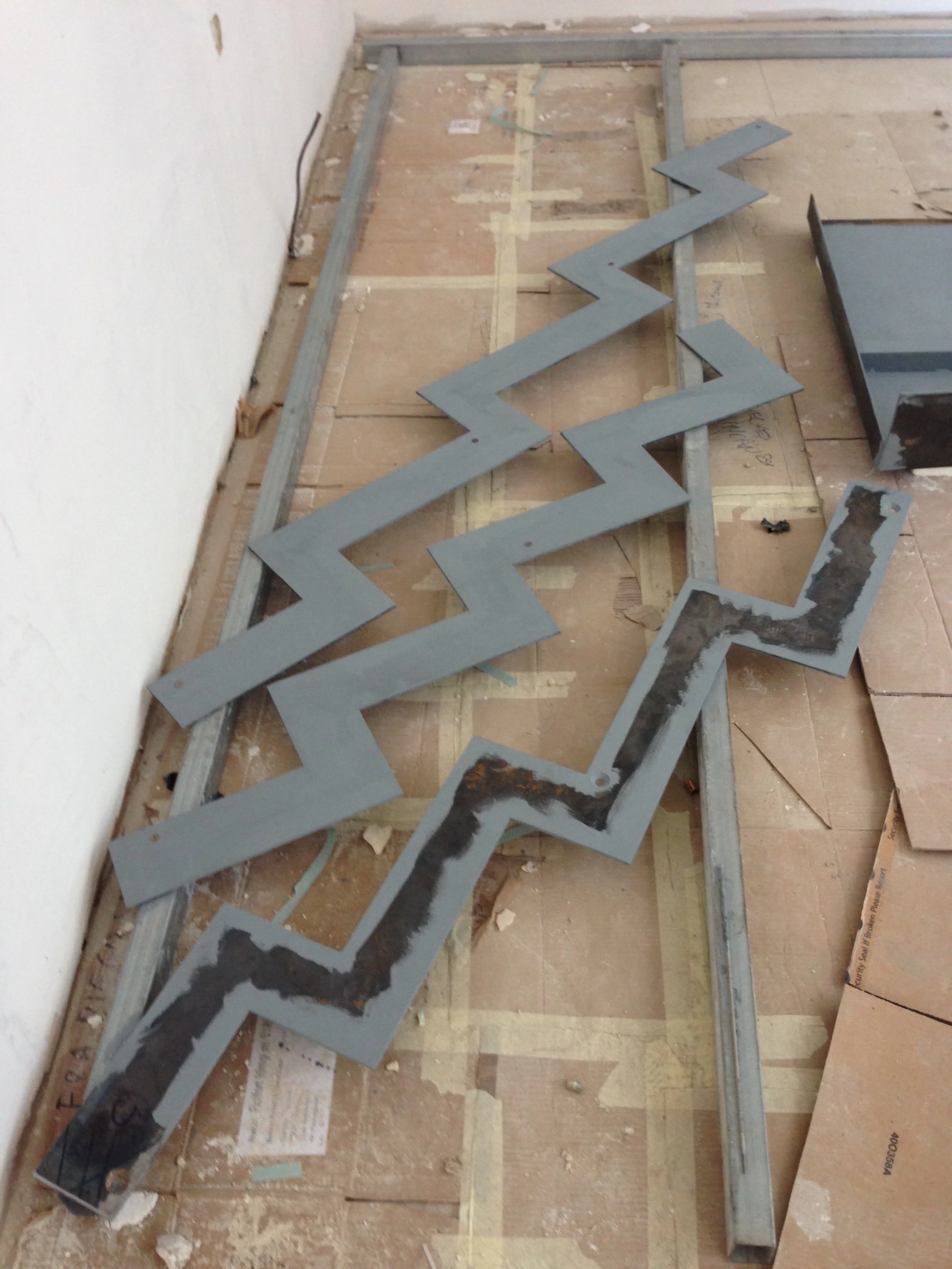 Construction site with a metal zigzag-shaped piece leaning against a boilerplate floor, drywall, and framing materials.