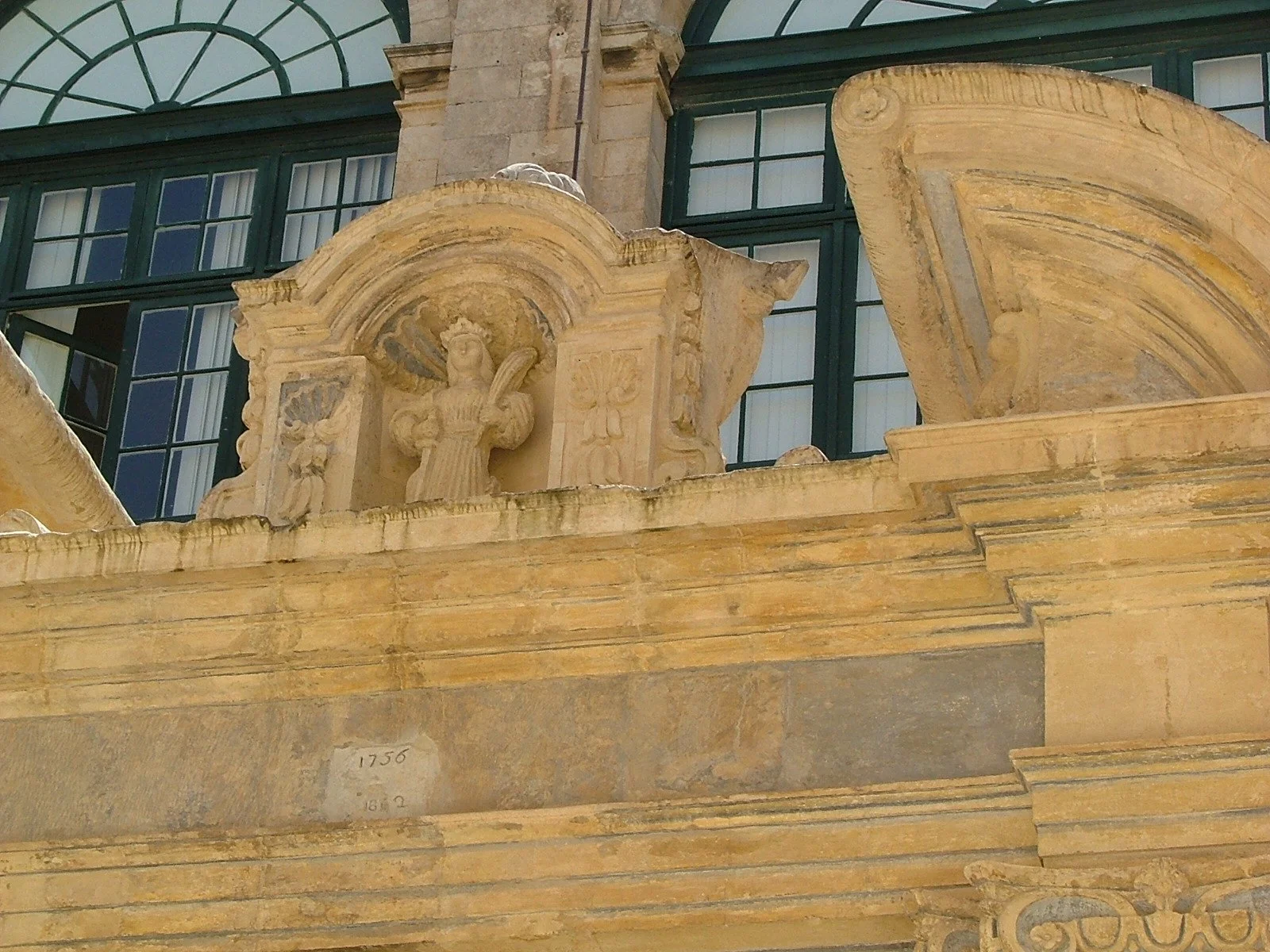 Close-up of an architectural detail on a historic building, featuring a carved stone bust of a person with a feather, set within an ornate arched niche, with large windows in the background.