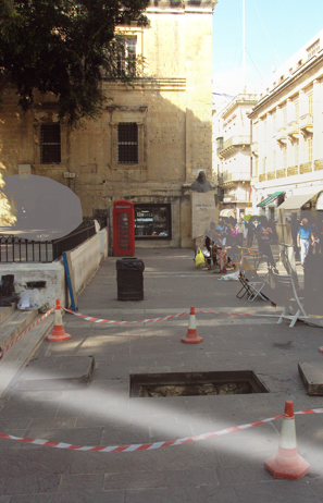 Street scene with construction area, orange cones, and a hole in the sidewalk, surrounded by buildings and pedestrians.