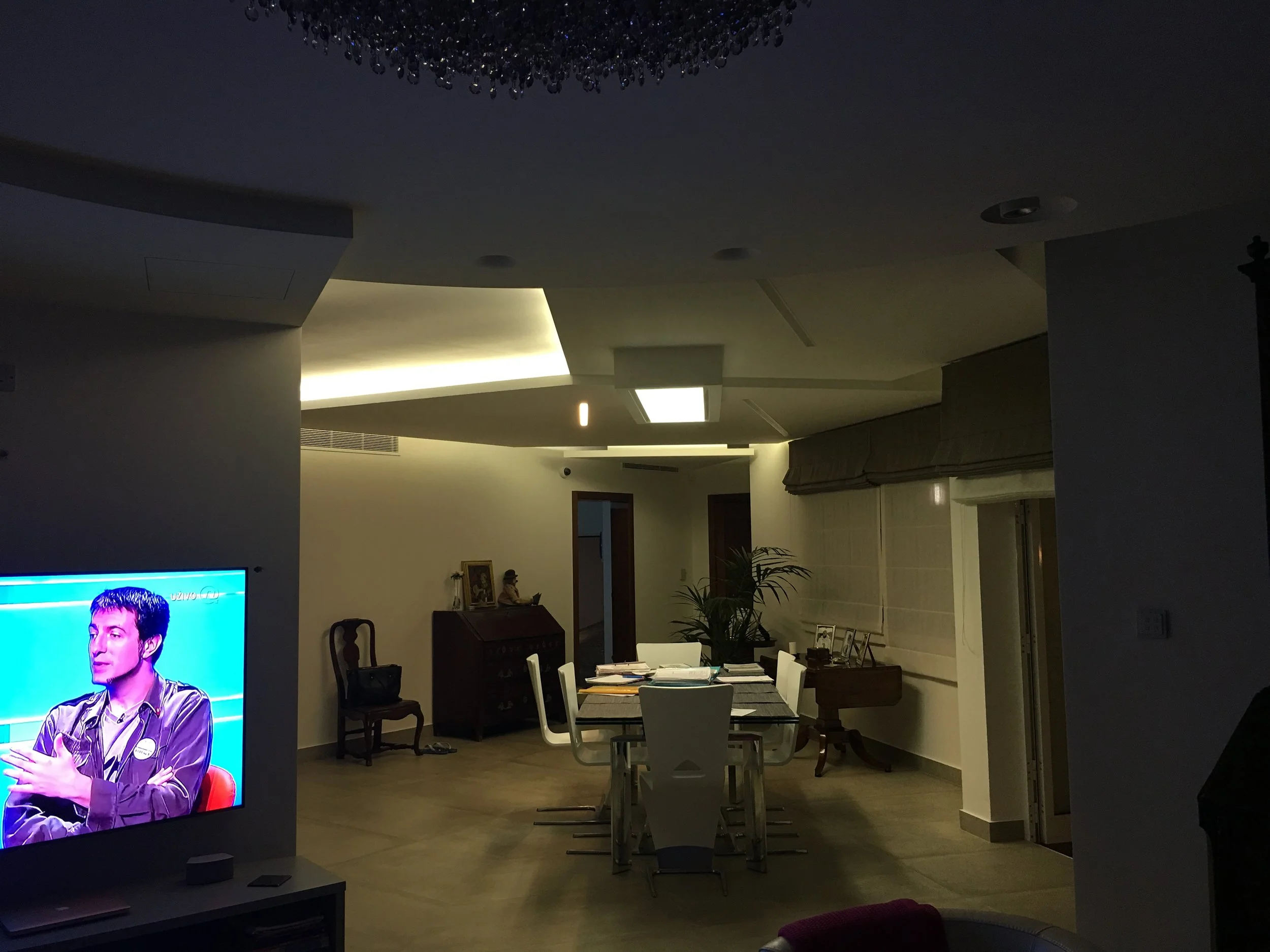 A dimly lit dining area with a modern design. There is a table surrounded by chairs, with a few items and papers on the table. In the background, a chandelier hangs from the ceiling. To the left, there is a large television screen showing a man in a 