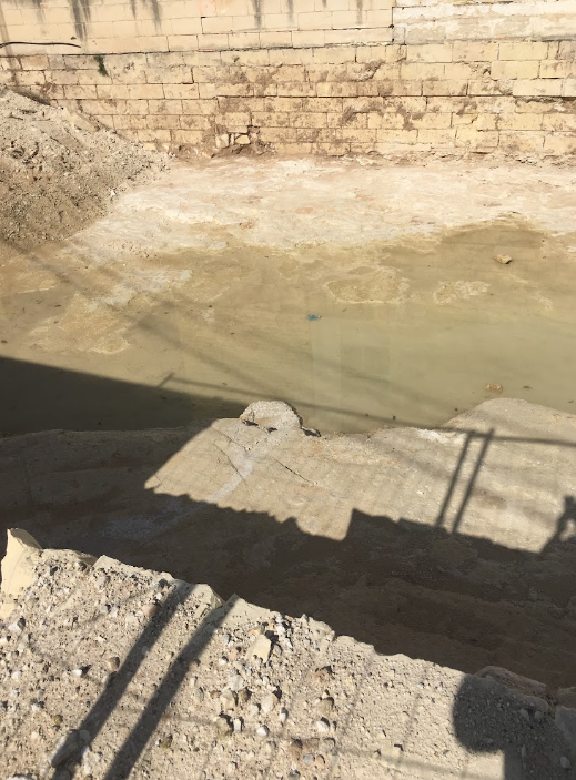 Empty construction site with a partially built brick wall and a large hole filled with water.