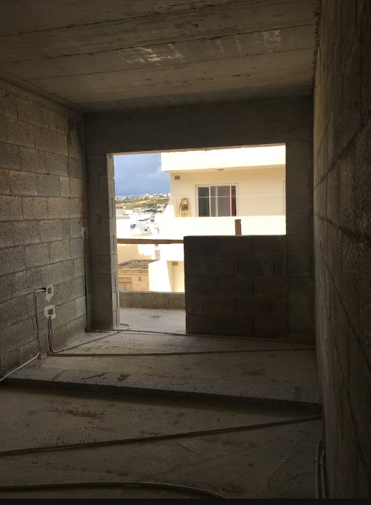 View from inside an unfinished concrete building looking out through a window opening at neighboring buildings with a cloudy sky.