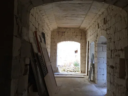 View of an arched stone tunnel with construction materials leaning against the wall on the left side and a window at the end showing an outdoor scene.