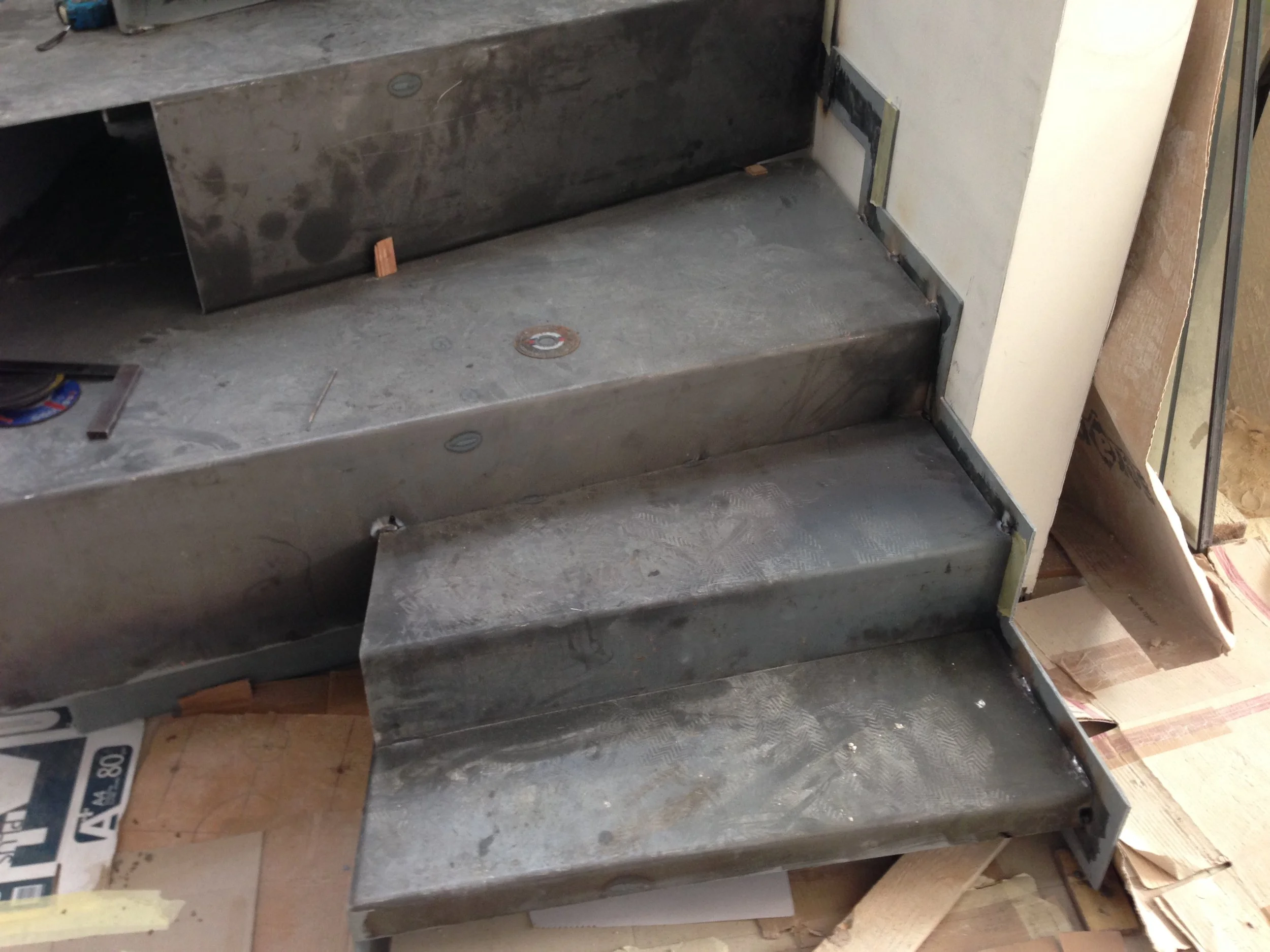 Metal staircase with four steps, visible screws, some dirt, and wooden blocks near the bottom step, adjacent to a wall and some construction materials.