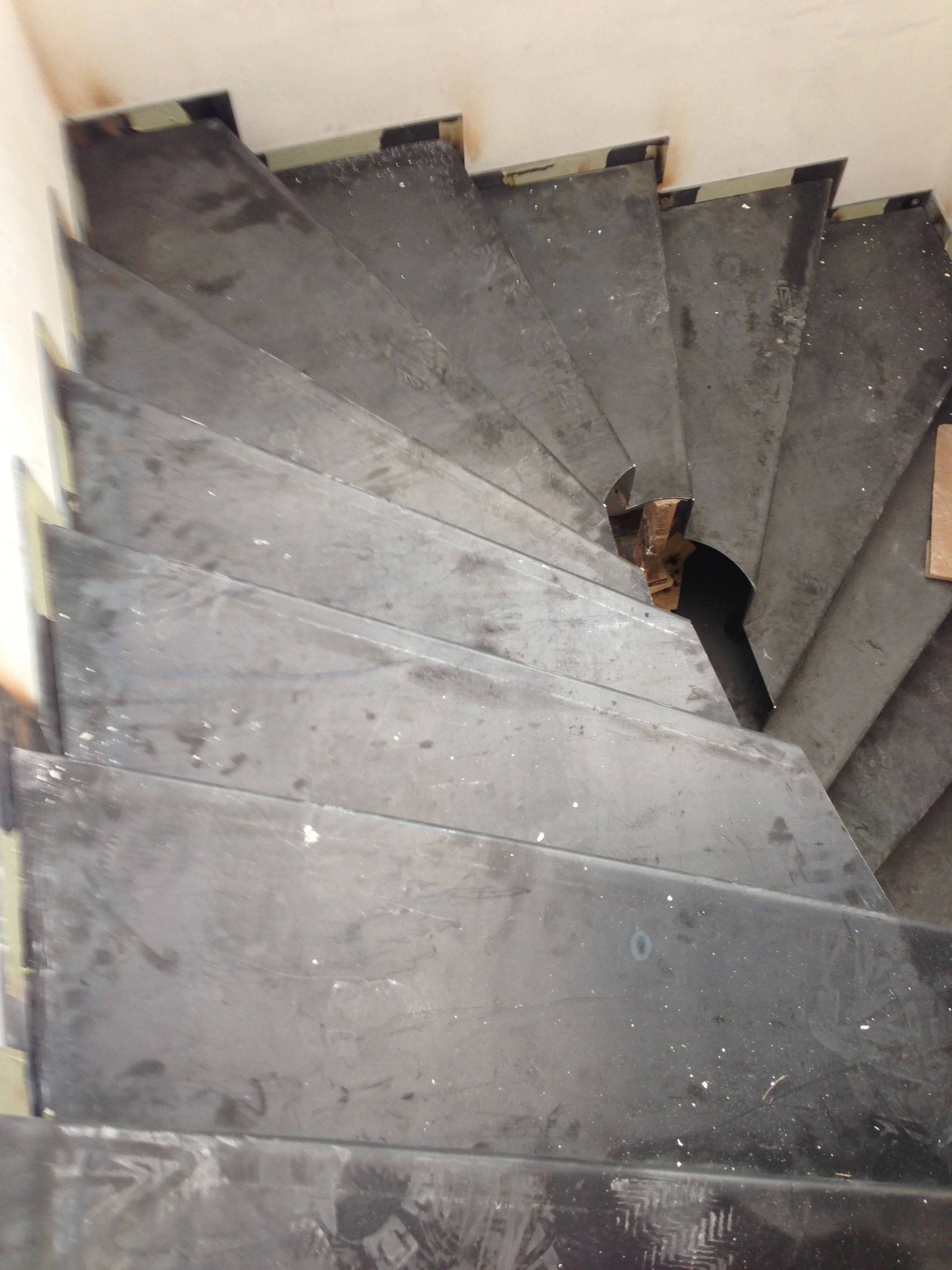 Metal staircase with black steps leading downwards, adjacent to a beige padded wall, with visible screws and some dirt or debris on the steps.