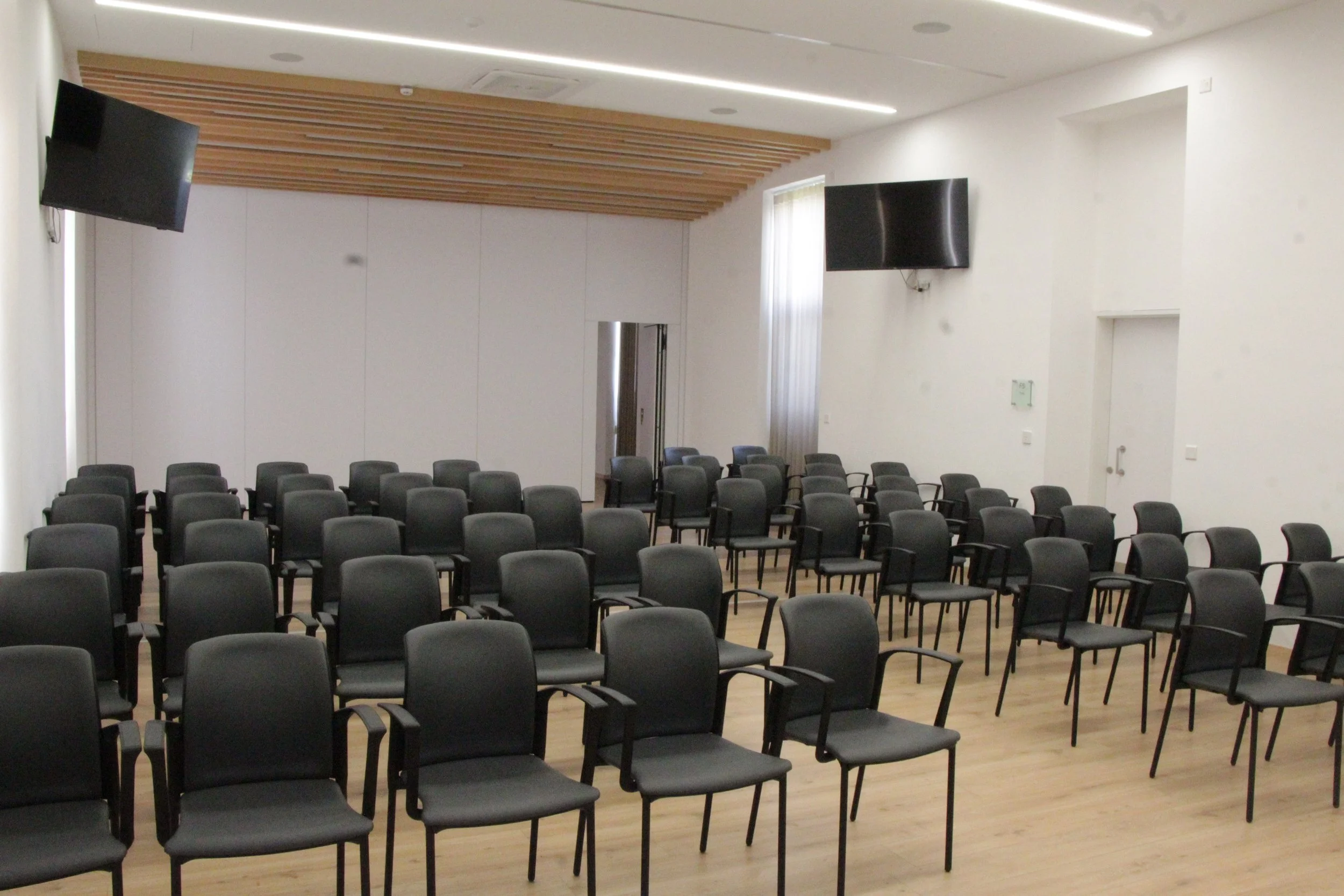An empty conference or lecture room with multiple rows of black chairs, two large flat-screen TVs mounted on the walls, white walls, a ceiling with recessed lighting, a wooden accent on the ceiling, a vertical window with curtains, and a door.