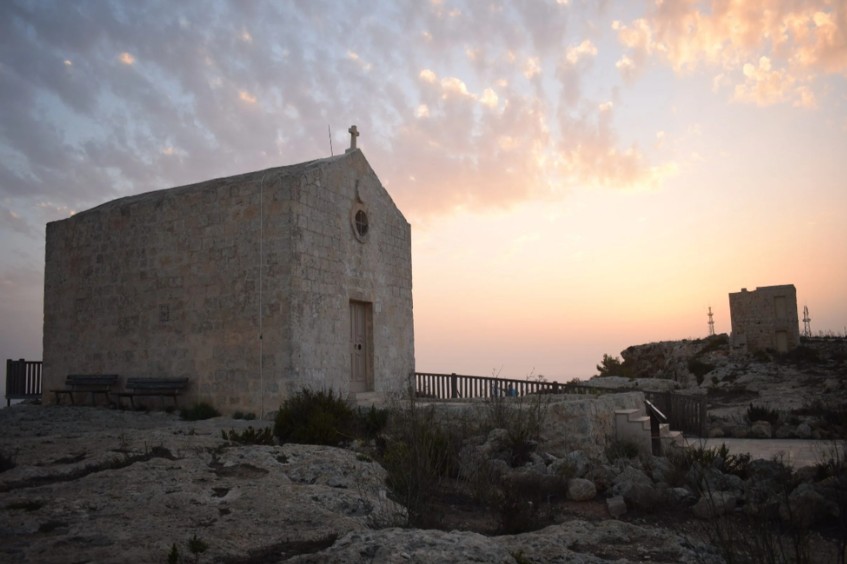 Small stone church with a cross on top, benches outside, and a sunset sky with scattered clouds in the background.