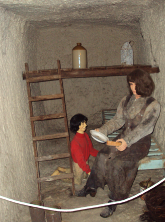 Wax figure scene of a woman sitting on a bench handing a plate to a young boy in a small, rustic room with a wooden ladder and a jar on a shelf.