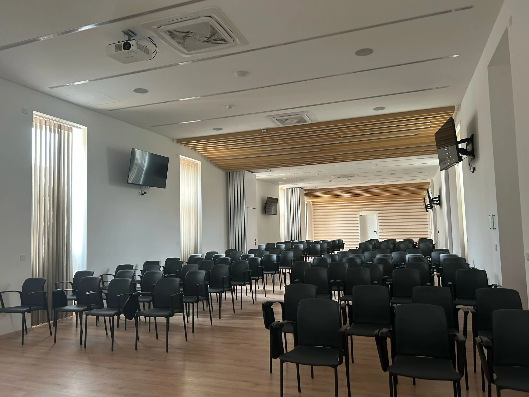 An empty conference room with rows of black chairs, large windows with vertical blinds, and wall-mounted flat-screen TVs.
