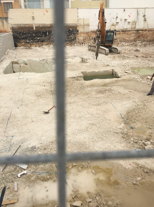 Construction site with excavator and dug holes in the ground, viewed through a chain-link fence.