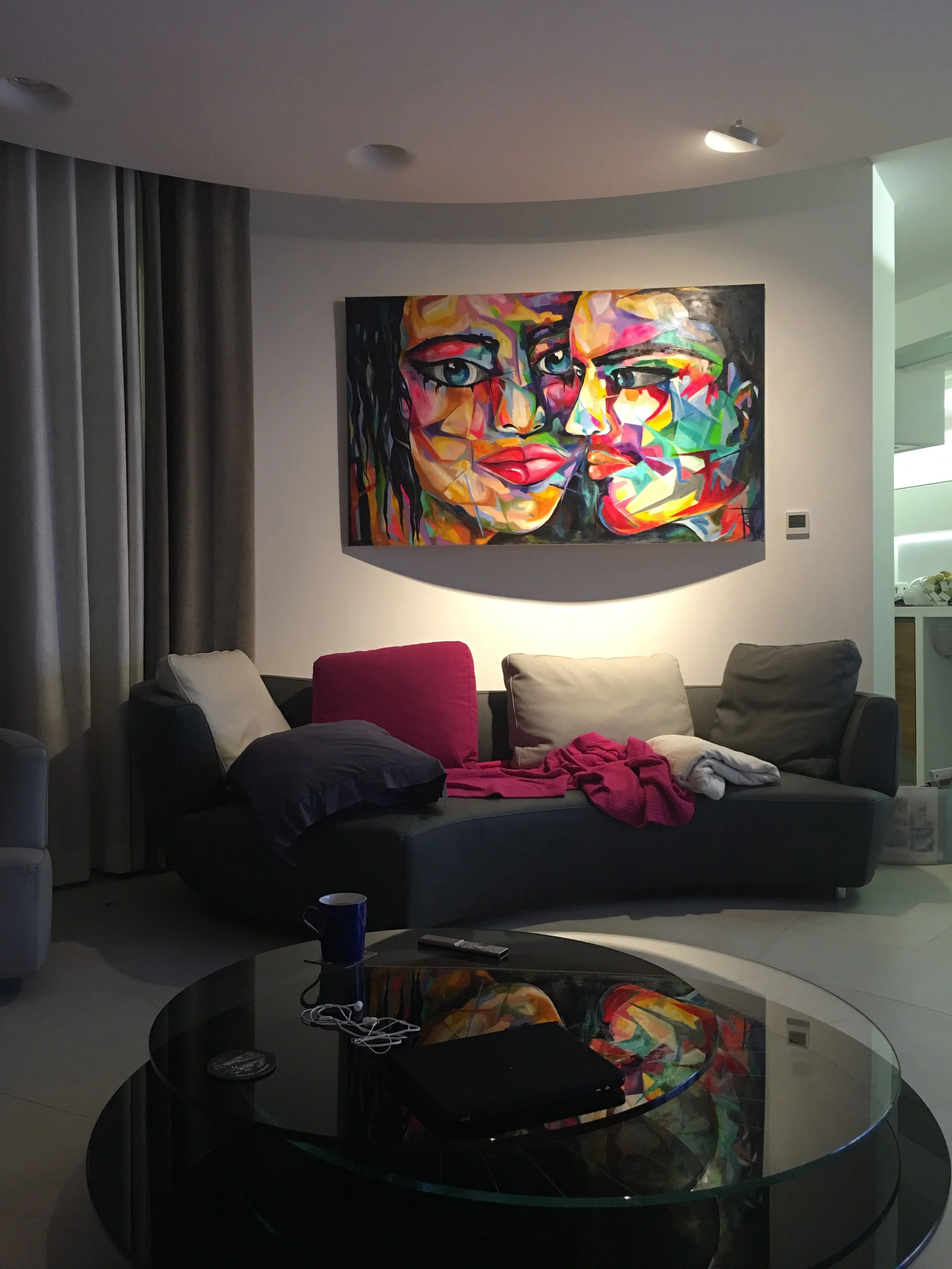 A brightly colored abstract painting of human faces on the wall above a dark gray sofa with multiple cushions, in a living room with a glass coffee table, some clothing, a remote, a cup, and a screen remote control.