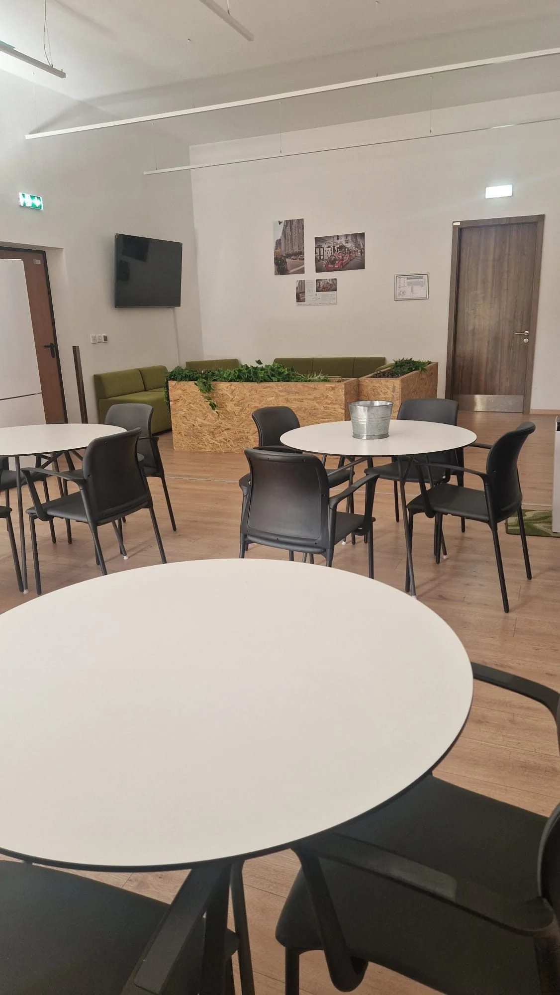 A modern dining area with round white tables, black chairs, a green cushioned bench, and potted plants in a wooden planter. There is a TV on the wall and framed pictures, with a wooden door and white walls.