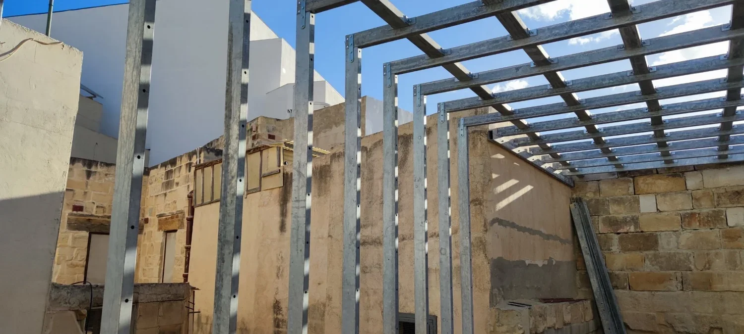 Metal framing for a building under construction with partially built brick and concrete walls and a blue sky.